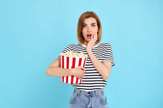 scared woman watching horror movie holding bucket of popcorn isolated on blue background