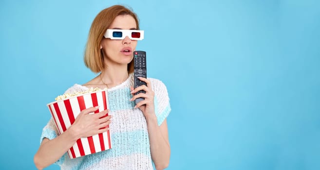 beautiful woman in 3d glasses with remote control enjoying interesting movie and holding bucket of popcorn on blue background. copy space