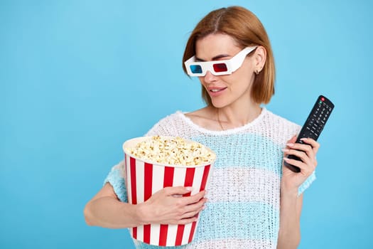 beautiful woman in 3d glasses with remote control watching movie film and holding bucket of popcorn on blue background. copy space
