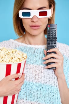 beautiful woman in 3d glasses using tv control, watching movie film and holding bucket of popcorn on blue background