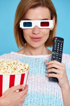 beautiful woman in 3d glasses with remote control holding bucket of popcorn on blue background