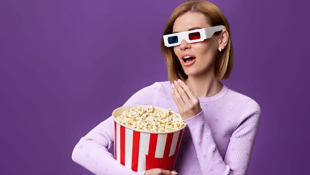 jouful caucasian woman in 3d glasses watching movie film, eating popcorn isolated on purple background