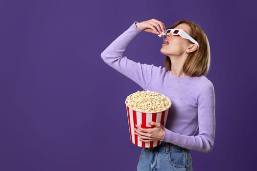 jouful caucasian woman in 3d glasses watching movie film, eating popcorn isolated on purple background