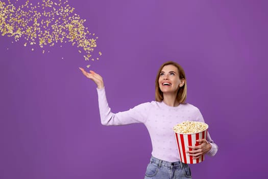 happy beautiful woman holding bucket with flying popcorn isolated on purple background