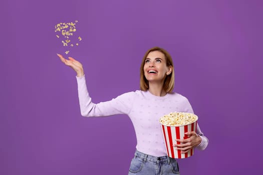 happy beautiful woman holding bucket with flying popcorn isolated on purple background