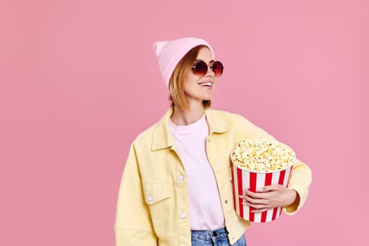 Cheerful smiling woman in sunglasses holding bucket of popcorn isolated on pastel pink background.