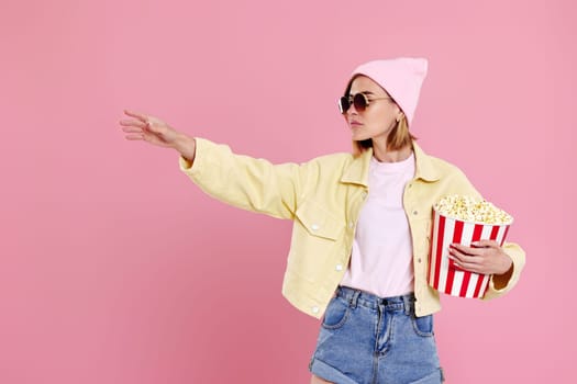 Cheerful young woman in sunglasses, hat with bucket of popcorn pointing at copy space on pastel pink background.
