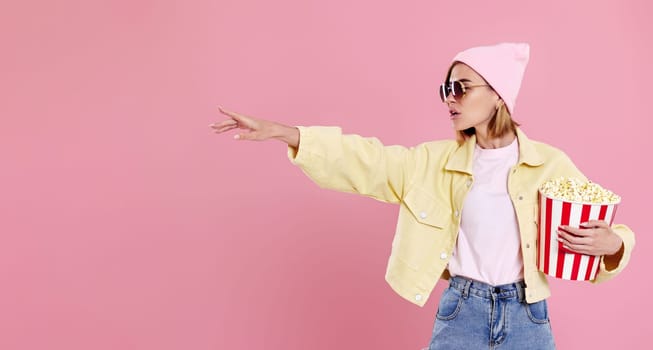 Cheerful young woman in sunglasses, hat with bucket of popcorn pointing at copy space on pastel pink background.