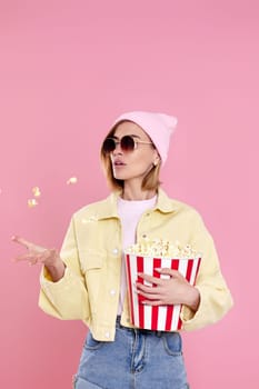 Cheerful happy woman in sunglasses, hat holding bucket of popcorn isolated on pastel pink background.