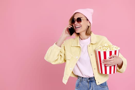 young beautiful woman in sunglasses, hat with bucket of popcorn talking on the phone on pastel pink background. copy space