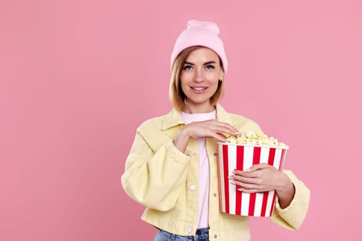 happy girl in hat watching movie film, holding bucket of popcorn isolated on pastel pink background.