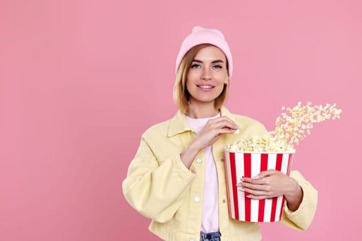 beautiful young woman in hat watching movie film, holding bucket of popcorn isolated on pastel pink background.