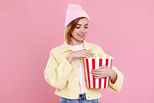 Cheerful young woman in hat watching movie film, holding bucket of popcorn on pastel pink background.