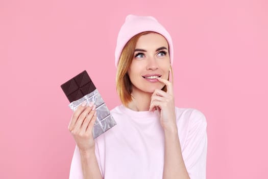 Stylish young blonde woman in t-shirt and hat holding chocolate bar on pink background