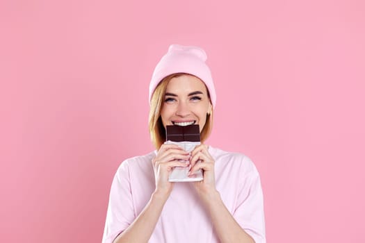 Beautiful young woman in casual t-shirt biting delicious chocolate bar and looking at camera isolated on pastel pink background