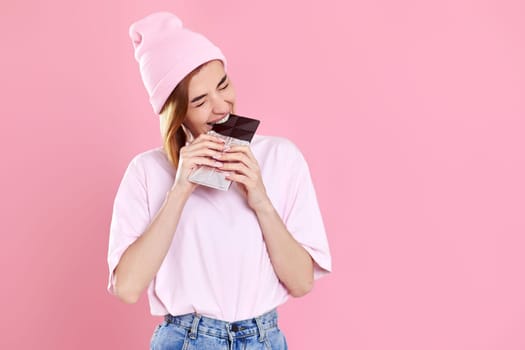 charming woman in casual t-shirt biting sweet chocolate bar isolated on pastel pink background