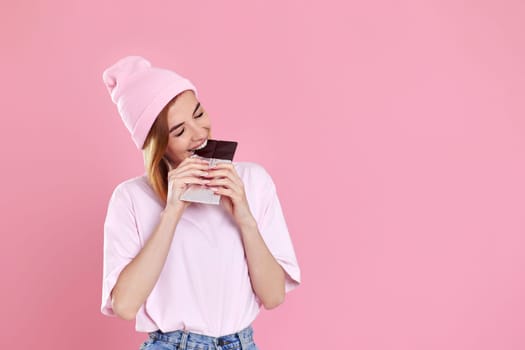 Beautiful young woman in casual t-shirt biting delicious chocolate bar isolated on pastel pink background