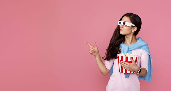 Cheerful young woman in hat with bucket of popcorn pointing at copy space on pink background.