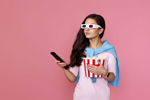 pretty woman in 3d glasses with remote control holding bucket of popcorn on pink background. copy space