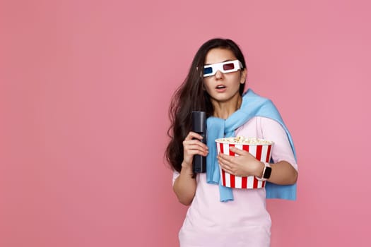 scared terrified woman in 3d glasses with remote control enjoying interesting movie and holding bucket of popcorn on pink background. copy space