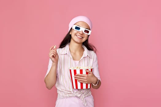 smiling caucasian girl in pink hat watching movie film, holding bucket of popcorn isolated on pastel pink background.