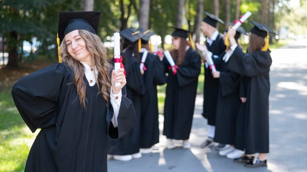 Group of happy students in graduation gowns outdoors. A young girl with a diploma in her hands in the foreground