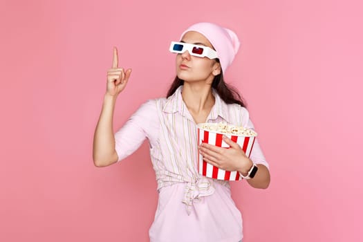 Cheerful young woman in hat with bucket of popcorn pointing up at copy space on pink background.