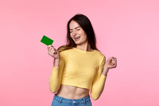 amazed glad woman in yellow clothes holding plastic credit card isolated on pink background