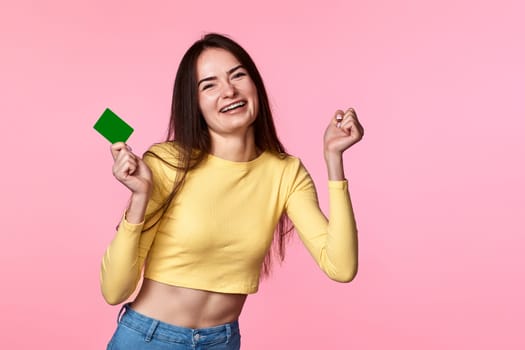 young Joyful woman in yellow clothes holding plastic credit card isolated on pink background