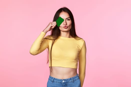 young woman covering eye with plastic credit card isolated on pink background