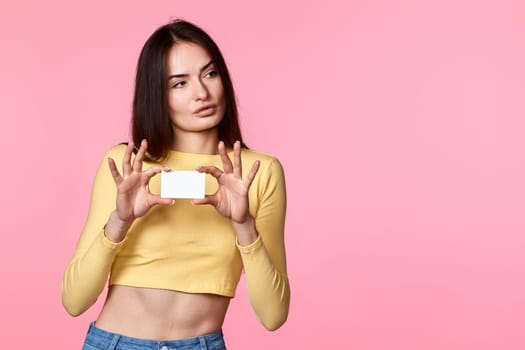 young woman in yellow clothes holding plastic credit card and looking at empty copy space isolated on pink background