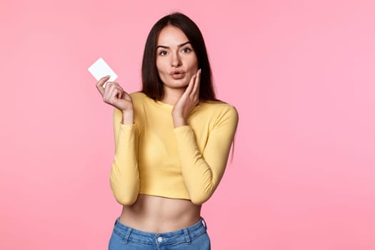 amazed surprised woman in yellow clothes holding plastic credit card isolated on pink background