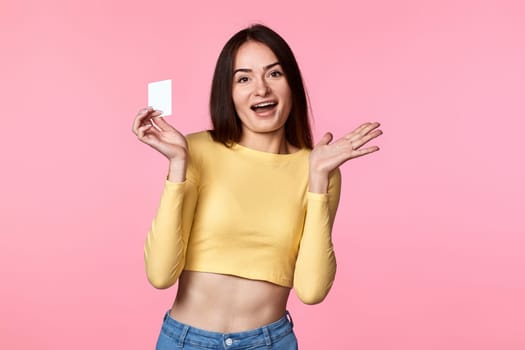 amazed glad woman in yellow clothes holding plastic credit card isolated on pink background