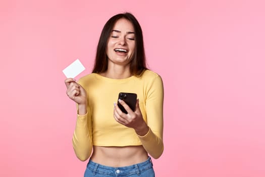 happy attractive woman paying with plastic credit card on smartphone app isolated on pink background. Online payment