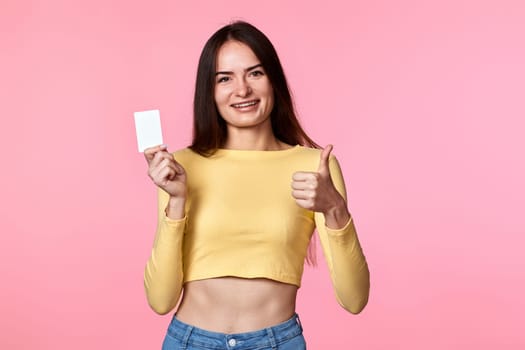 young woman in yellow clothes holding plastic credit card isolated on pink background