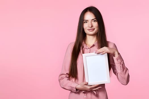 adorable woman in pink dress holding white photo frame on pink background. copy space