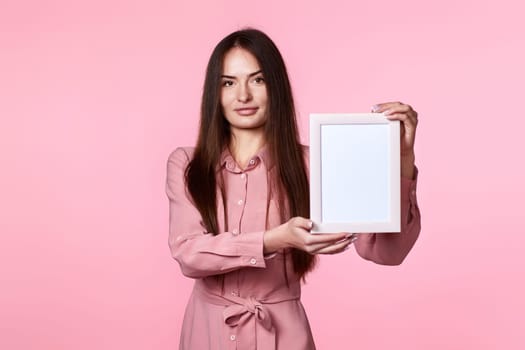 young charming woman in pink dress holding white photo frame on pink background