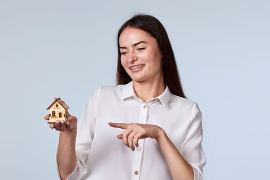 friendly smiling woman pointing finger at little house on gray background. real estate insurance concept