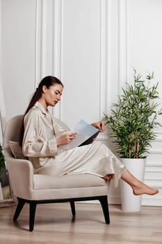 brunette woman in silk beige pajamas sits in chair and reads magazine in room