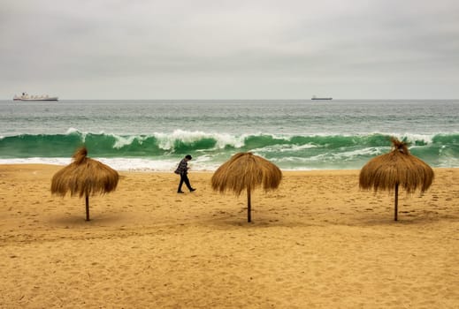 Single man walking past three palm umbrellas on an empty beach in Valparaiso Chile on a cold summer day