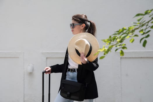 young happy brunette girl in sunglasses and straw hat with a suitcase goes on vacation, a woman stands against the background of a white brick wall, High quality photo