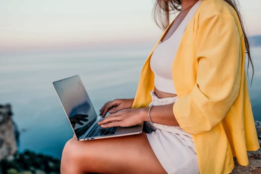 Successful business woman in yellow hat working on laptop by the sea. Pretty lady typing on computer at summer day outdoors. Freelance, travel and holidays concept.