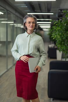 Attractive mature caucasian woman holding laptop while standing in office