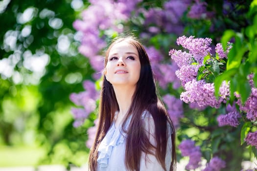 A beautiful happy girl stands next to a flowering lilac bush in the park, looks up Close up. Copy space