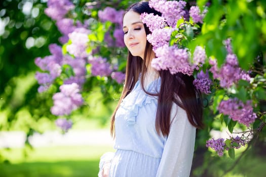 A beautiful girl in a light blue dress stands drowning in a flowering lilac bush in the park. Closing your eyes. Close up. Copy space