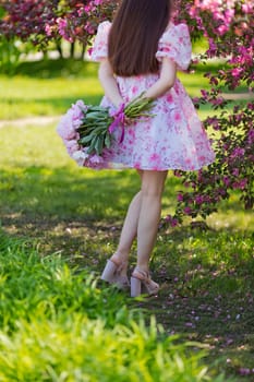 a girl with long hair, in a pink dress, standing with a large bouquet of peonies on green grass, in a blooming garden. Back view. Copy space