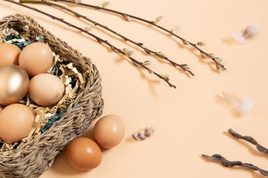 Easter eggs natural and gold in basket with paper filler . Branch of willow catkinsan feather. Light pastel orange background with copy space.