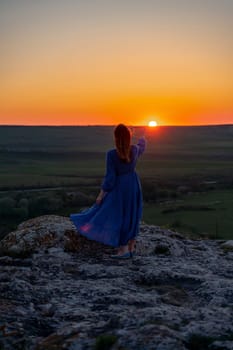Silhouette of a girl in a blue dress standing on a rock in the mountains at sunset. She held out her hand to the sun up