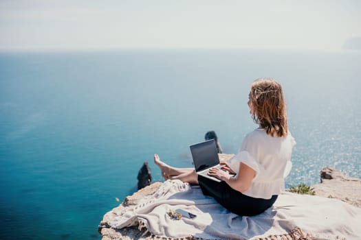 Successful business woman in yellow hat working on laptop by the sea. Pretty lady typing on computer at summer day outdoors. Freelance, travel and holidays concept.