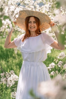 Woman cherry orchard. A happy woman in a long white long dress walks through the green spring blooming cherry garden. Happy cheerful princess bride. The fabric of the skirt flutters in the wind